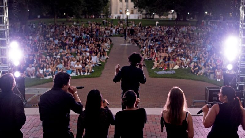 Concert pictured from behind facing the quad. People wearing black signing on stage and the crowd sitting on the ground watching.